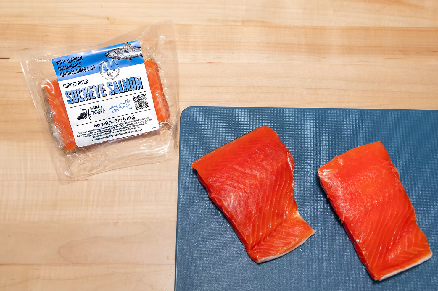 This image shows 3 Copper River Sockeye 6oz portions. 2 of them are out of the packaging on a blue cutting board, and the other one is in its packaging next to the cutting board.