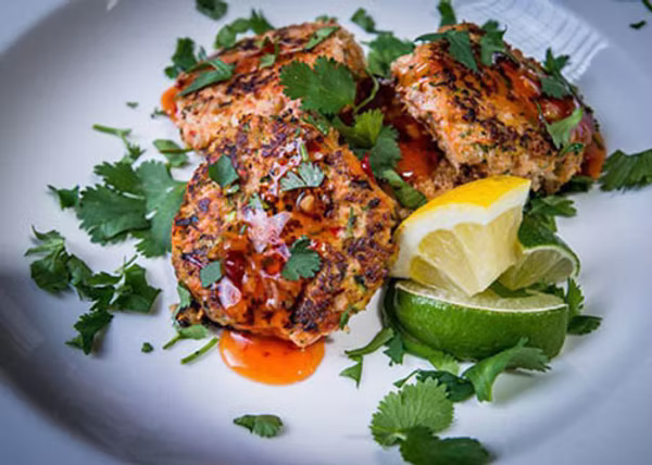 Minced sockeye salmon patties topped with cilantro, lemon, lime, and sweet chili sauce on a white plate.