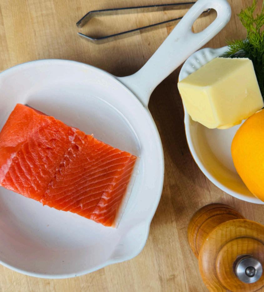 Raw salmon fillet in a white skillet with butter, an orange, and a pepper grinder on a wooden surface.