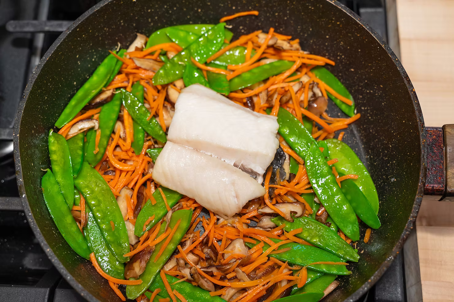 A 6 oz fillet of sablefish (black cod) atop a bed of peas, carrots and mushrooms in a saute pan.