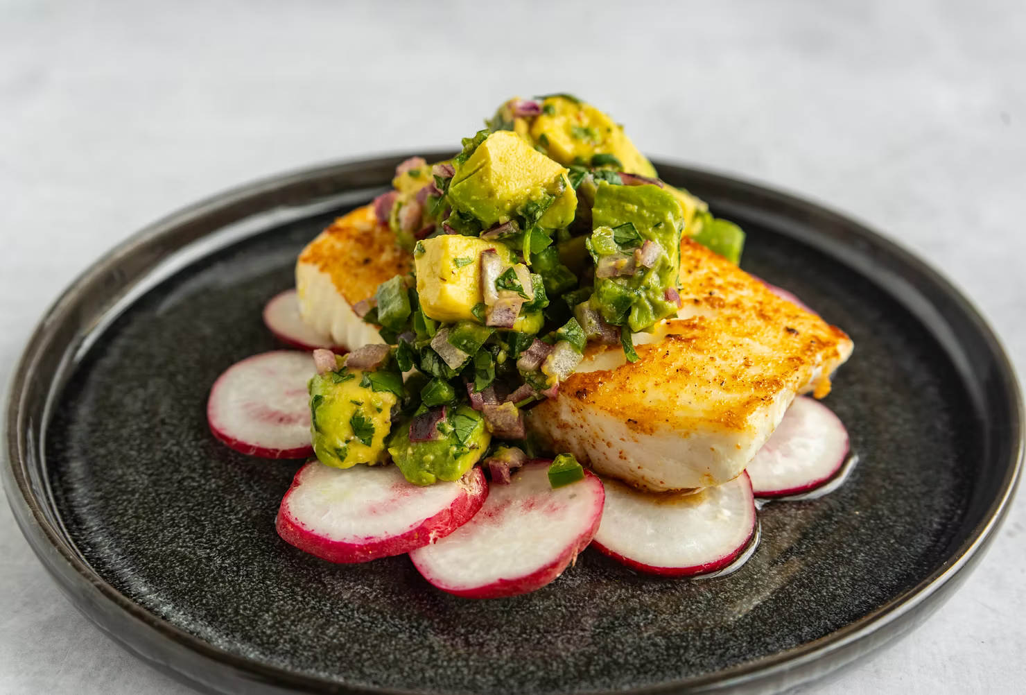 Halibut fillet topped with an avocado salsa over thinly sliced radishes on a black plate.