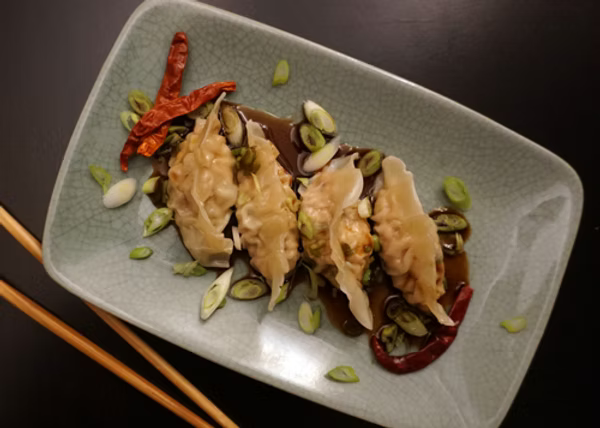 4 Copper River Salmon pot stickers plated over a dipping sauce and topped with green onions. They are on a green plate with chopsticks next to the plate on a black surface.