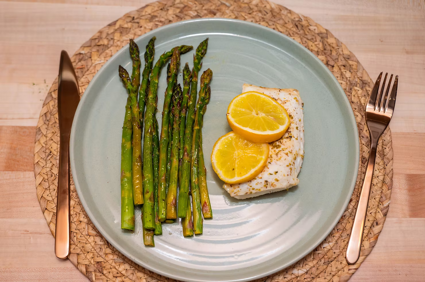 This image displays a piece of halibut topped with lemons, with asparagus on the side. It is on a blue plate with a knife on the left and a fork on the right.