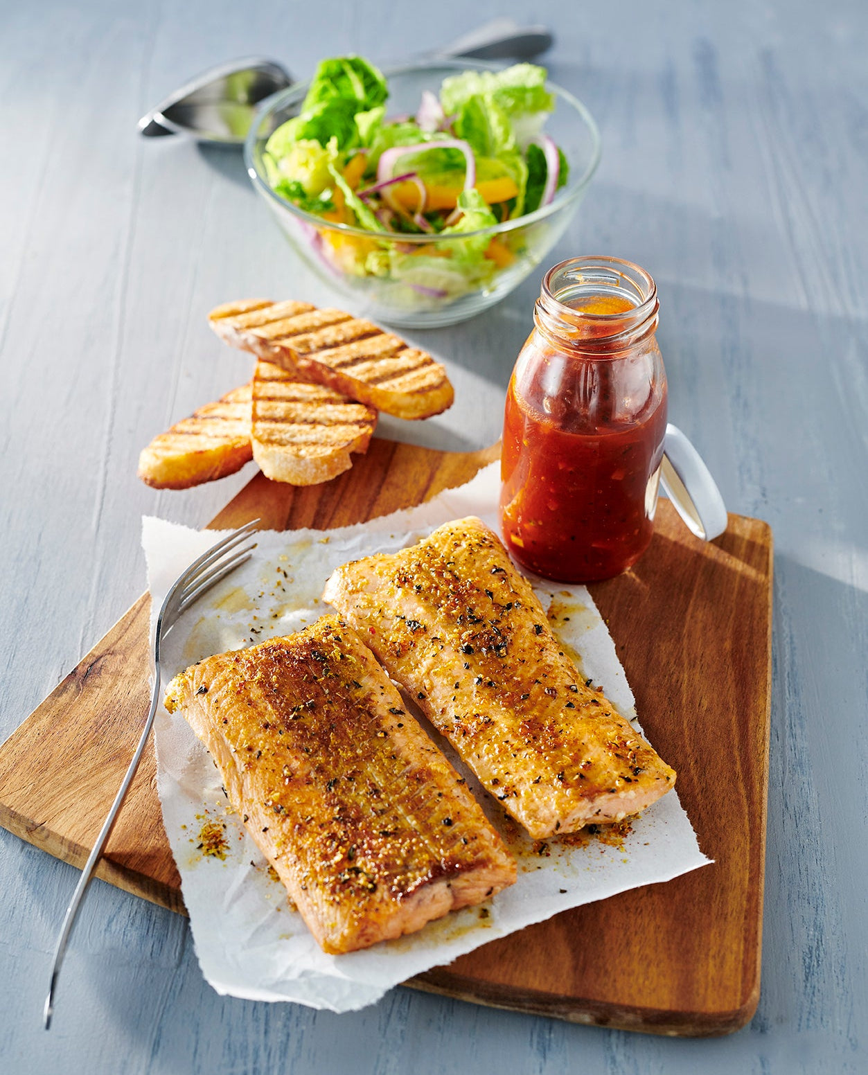 Grilled salmon fillets on a wooden board with a side of grilled toast, a small bottle of red sauce, and a salad bowl on a blue-gray wooden table.
