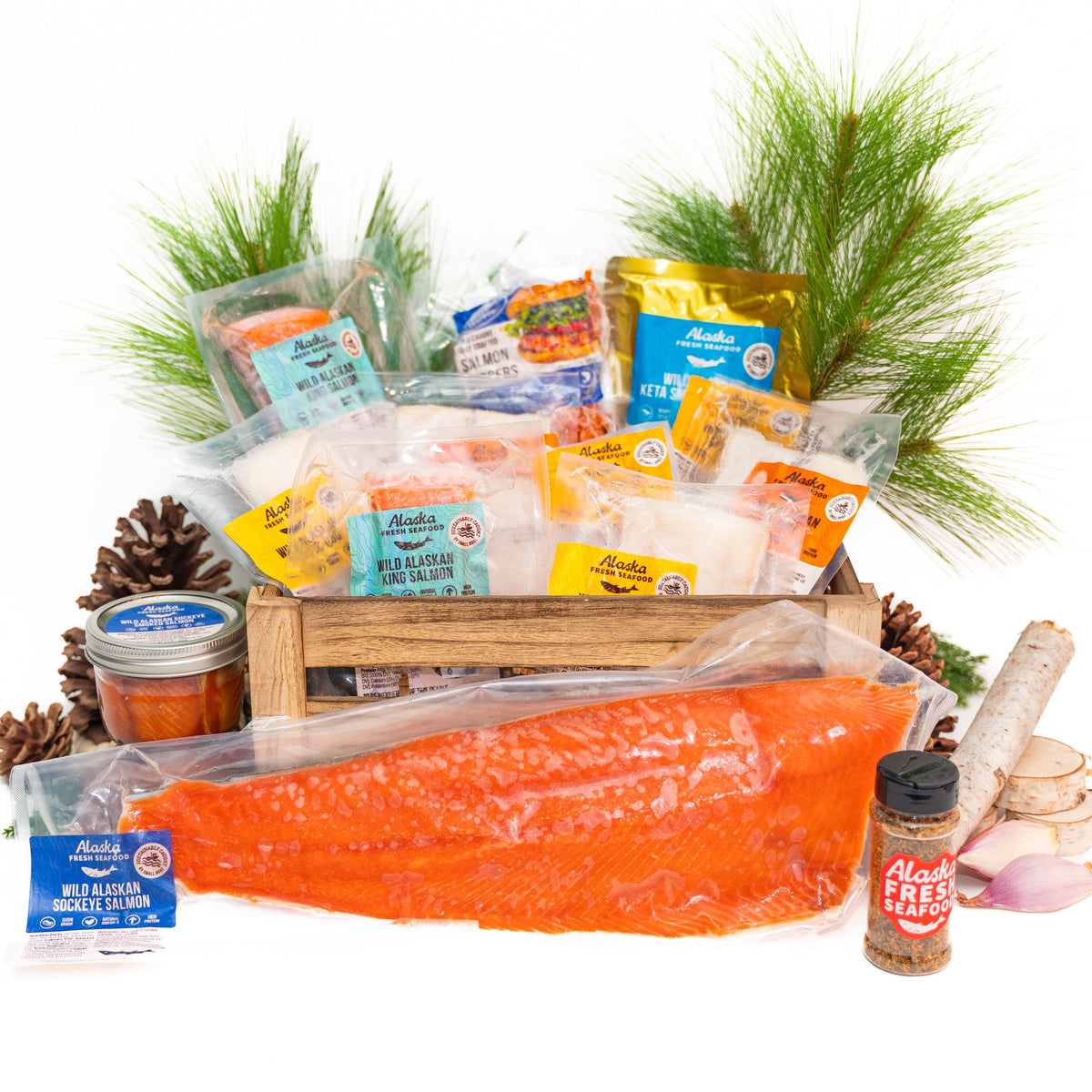 A Christmas-themed gourmet food gift basket arranged in a wooden crate on a white background, featuring a large orange salmon fillet, packaged meats, cheeses, spices, garlic, pine branches, and pinecones.