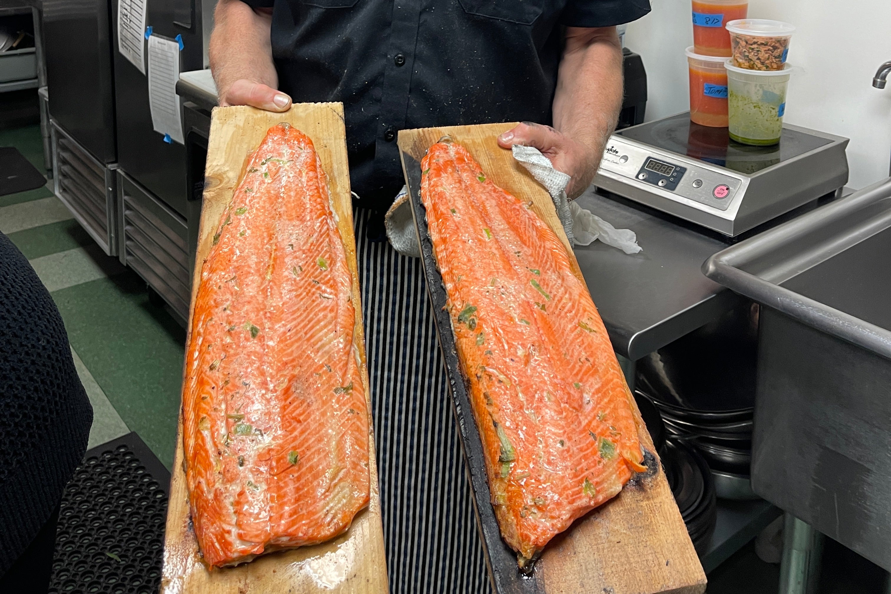 A smiling man in a black shirt stands in a commercial kitchen holding two large raw salmon fillets on wooden boards.