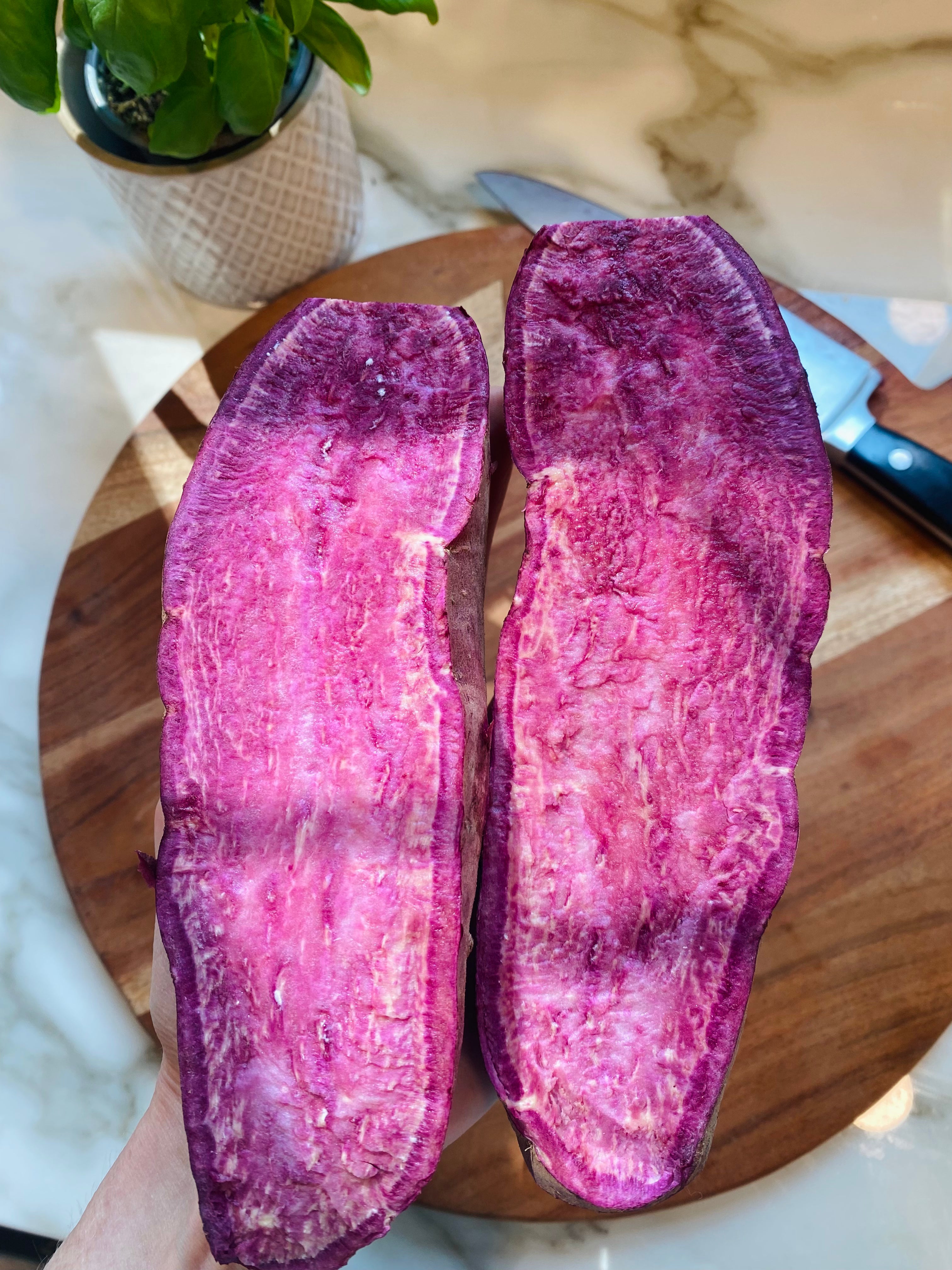 Two purple sweet potatoes on a wooden cutting board with a knife, on a light-colored surface.