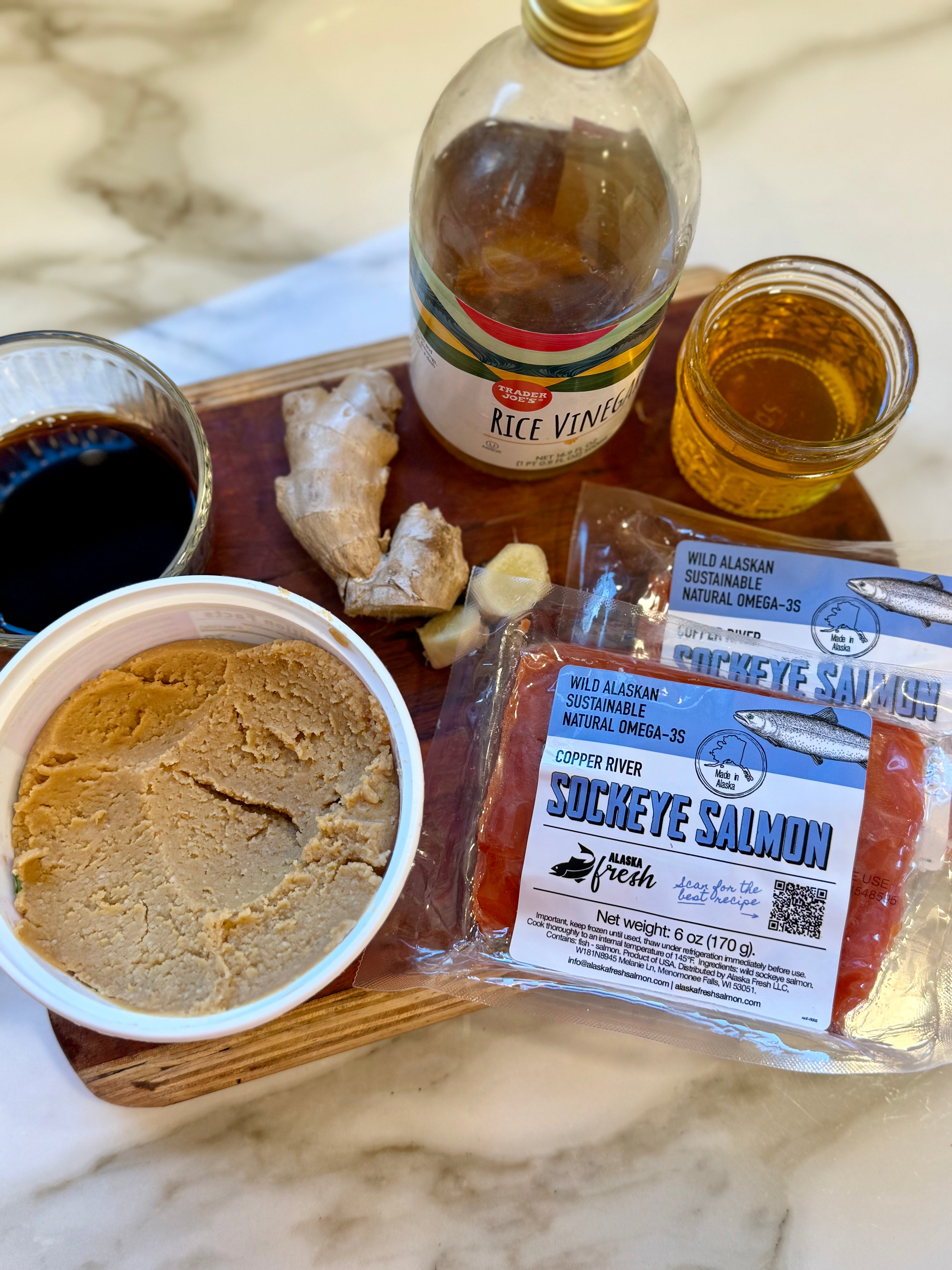 Assorted food items including a bottle of rice vinegar, a container of peanut butter, and a package of sockeye salmon on a marble surface.