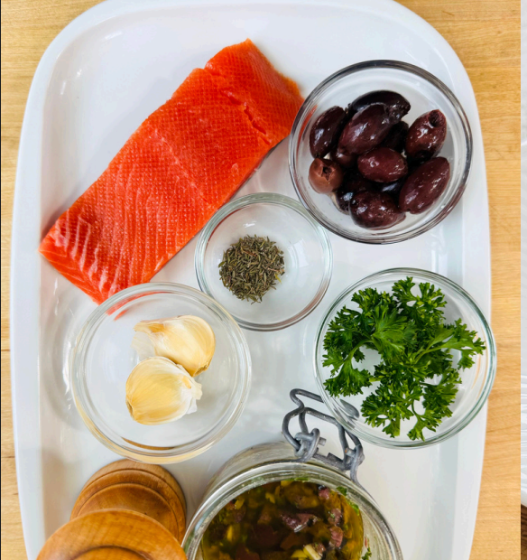 Salmon fillet with olives, herbs, and garlic on a white tray.