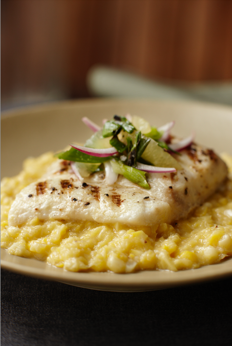 A close-up shot of a grilled fish fillet served on a bed of yellow risotto, topped with sliced red onion, green herbs, and a lemon wedge, presented on a beige plate.
